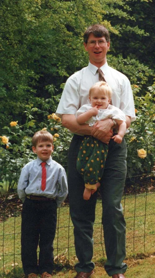 John Murphy standing in a garden holding a baby, with a small boy beside him