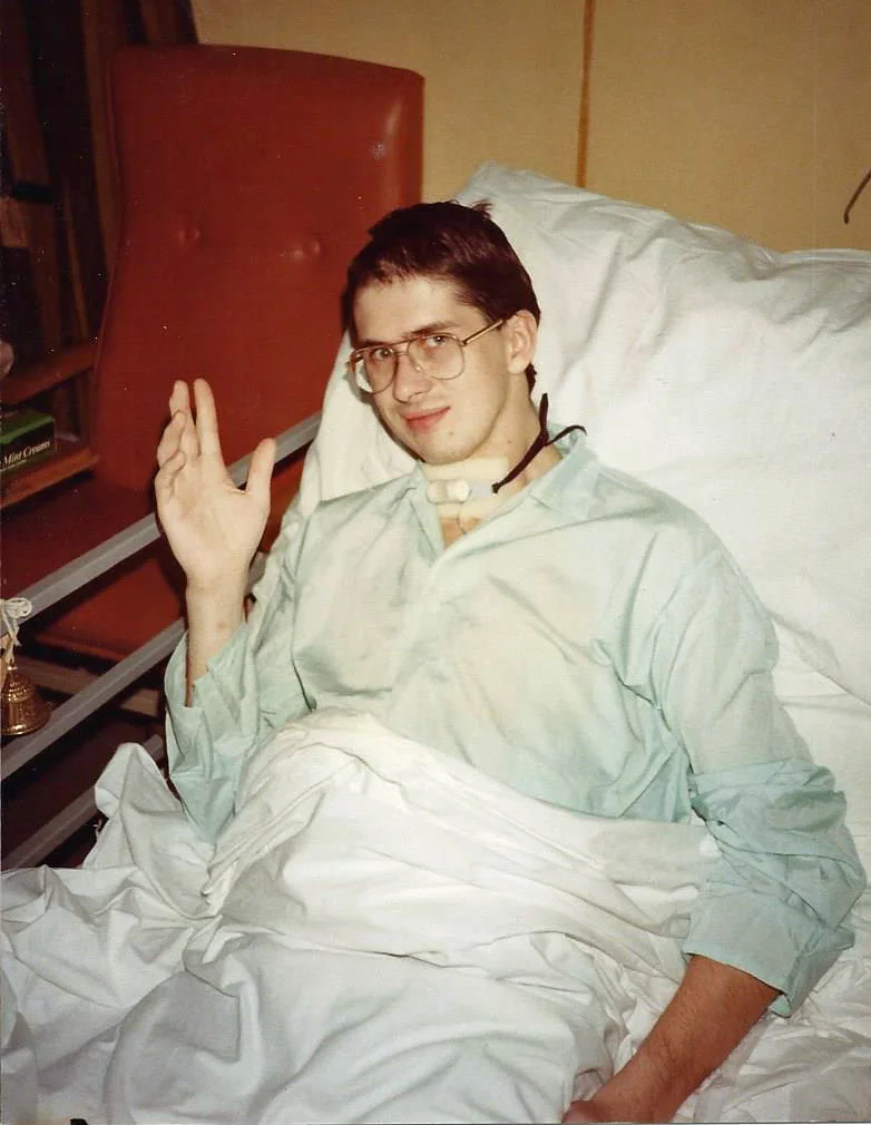 John Murphy sitting up in a hospital bed, raising a hand to wave